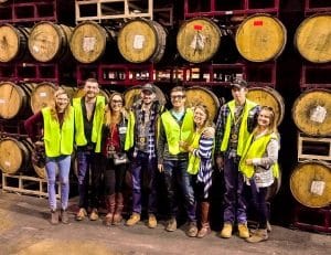 A group of Pittsburgh Brew Tour guests pose in front of stacks of barrels while wearing high visibility vests