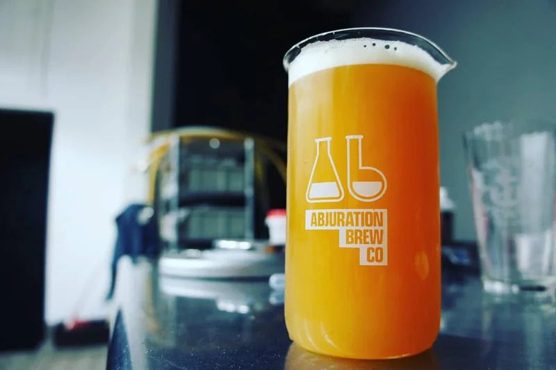 An Abjuration Brewing Co. branded beaker filled with a hazy beer.